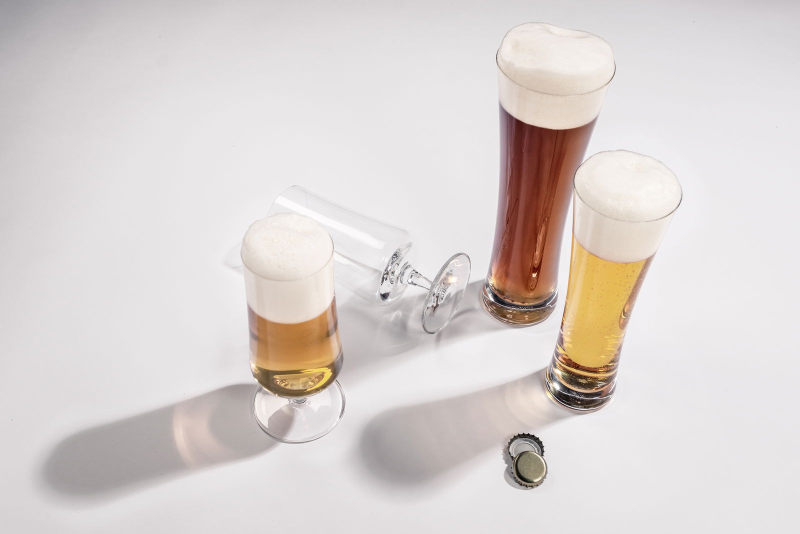 SCHOTT ZWIESEL 4-tlg. Weizenbiergläser Set BEER BASIC Draufsicht auf ein Set von vier Weizenbiergläsern, drei gefüllt mit Bier und Schaum, eines umgekippt, auf einem weißen Hintergrund mit zwei Kronkorken.