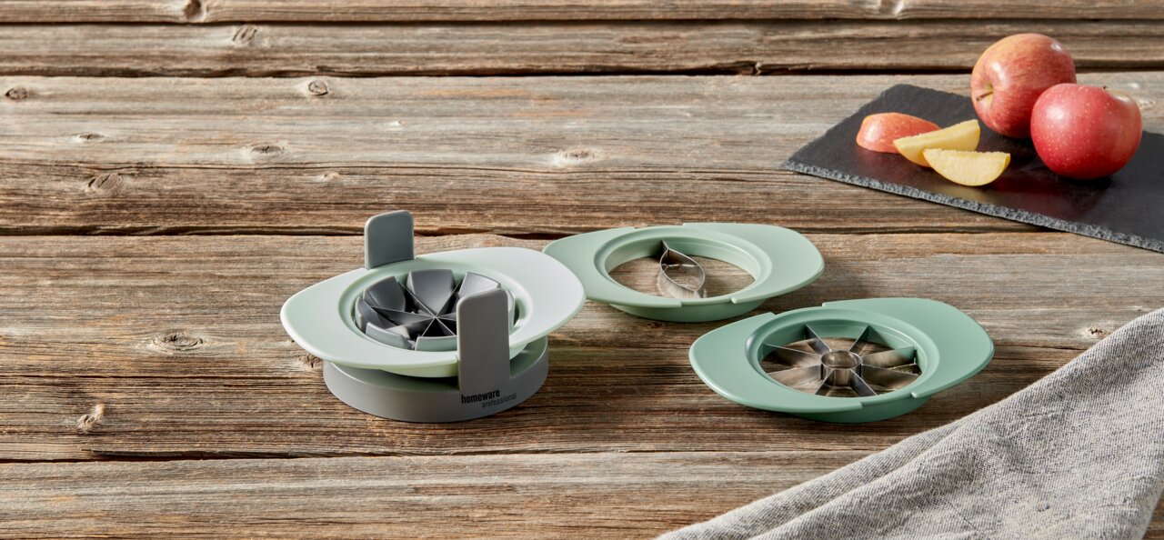 Homeware Apfelteiler 4er-Set PROFESSIONAL Apfelteiler in drei Teilen auf einem Holztisch, mit Äpfeln im Hintergrund, aus der Vogelperspektive fotografiert.