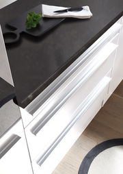 Moderne Küchenzeile mit weißem Hochglanz-Finish und dunkler Arbeitsplatte, von oben betrachtet. Auf der Arbeitsplatte liegt ein Schneidebrett mit einem Messer und Kräutern.