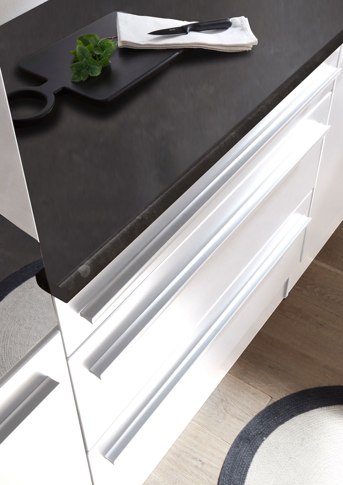 Moderne Küchenzeile mit weißem Hochglanz-Finish und dunkler Arbeitsplatte, von oben betrachtet. Auf der Arbeitsplatte liegt ein Schneidebrett mit einem Messer und Kräutern.