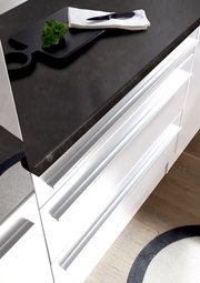 Moderne Küchenzeile mit weißem Hochglanz-Finish und dunkler Arbeitsplatte, von oben betrachtet. Auf der Arbeitsplatte liegt ein Schneidebrett mit einem Messer und Kräutern.