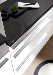 Moderne Küchenzeile mit weißem Hochglanz-Finish und dunkler Arbeitsplatte, von oben betrachtet. Auf der Arbeitsplatte liegt ein Schneidebrett mit einem Messer und Kräutern.