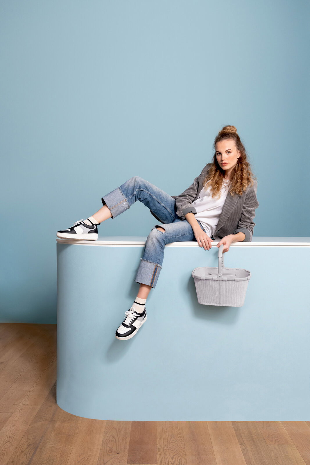 Junge Frau sitzt lässig auf einer hellblauen Theke und hält einen grauen Korb mit Henkel; Frontansicht in leicht seitlicher Perspektive.