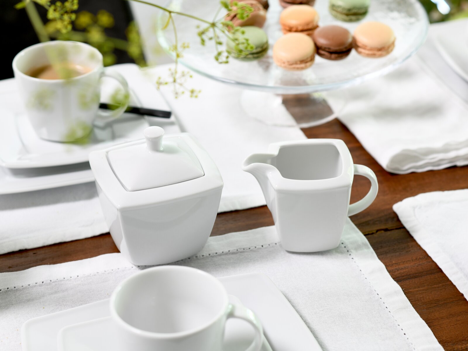 creaTable Milch und Zucker Set 2-tlg VICTORIA Weißes Porzellan-Set mit Zuckerdose und Milchkännchen auf gedecktem Tisch mit Tasse und Macarons im Hintergrund, leicht schräge Vorderansicht