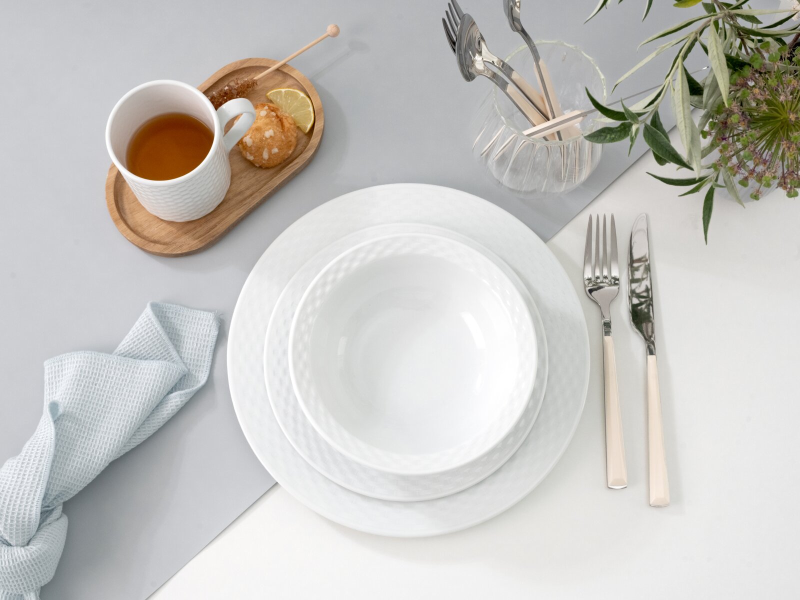 Drei weiße, strukturierte Porzellanteller im Set auf hellem Tischgedeck, mit Besteck und Tasse Tee; Perspektive: Draufsicht von oben