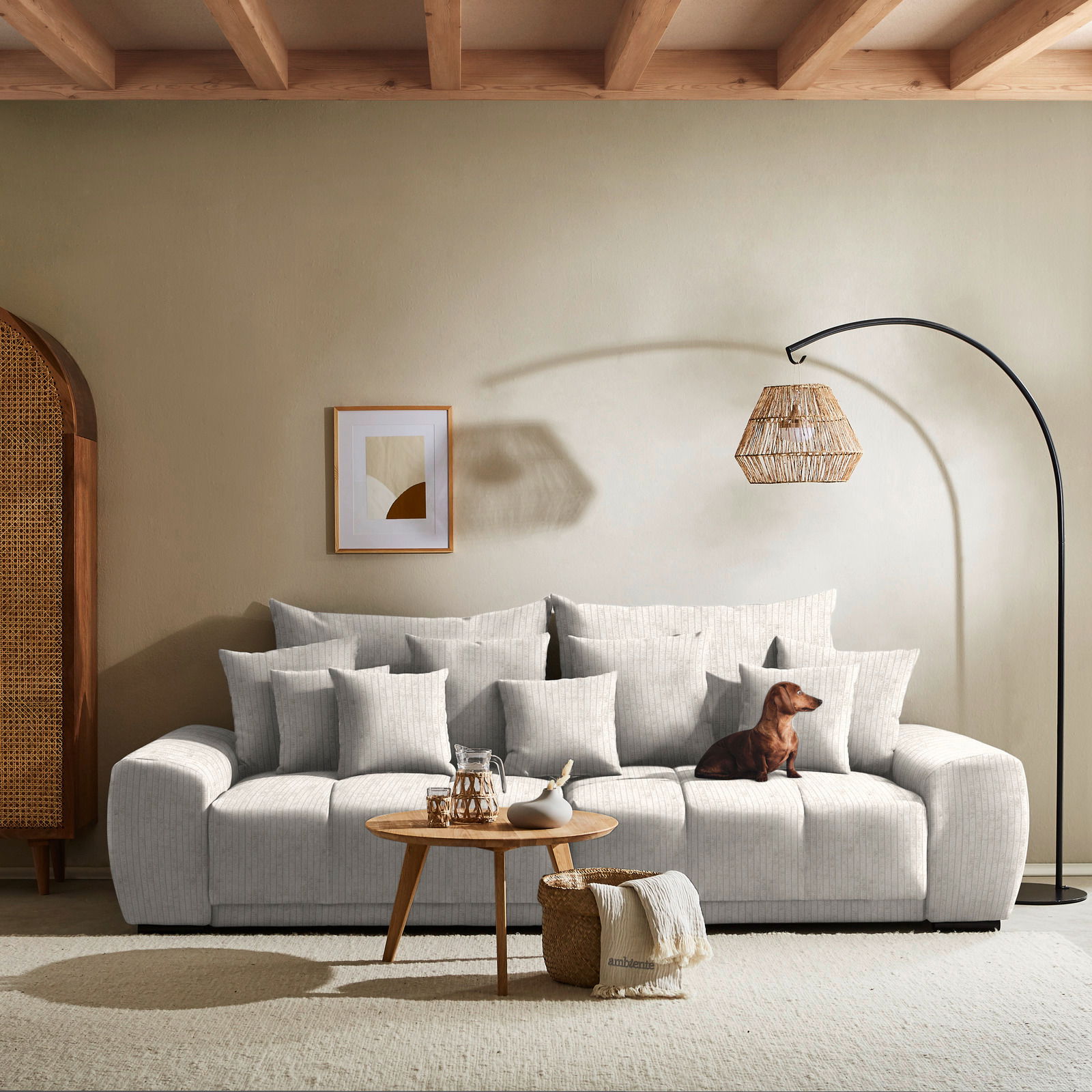 Großes, hellgraues Big-Sofa mit vielen Kissen, frontal fotografiert, mit einem kleinen Hund darauf und einem runden Holztisch davor.