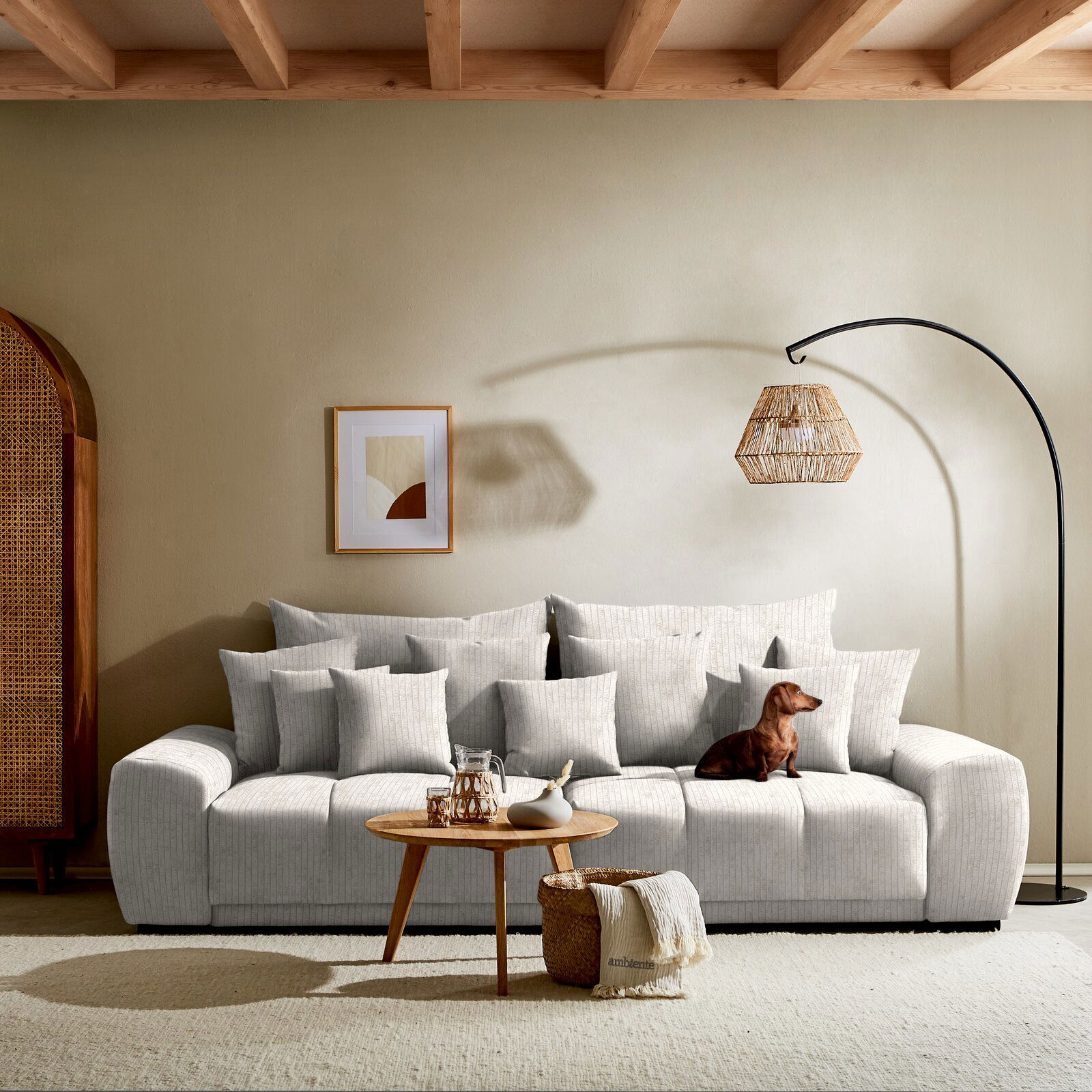 Großes, hellgraues Big-Sofa mit vielen Kissen, frontal fotografiert, mit einem kleinen Hund darauf und einem runden Holztisch davor.