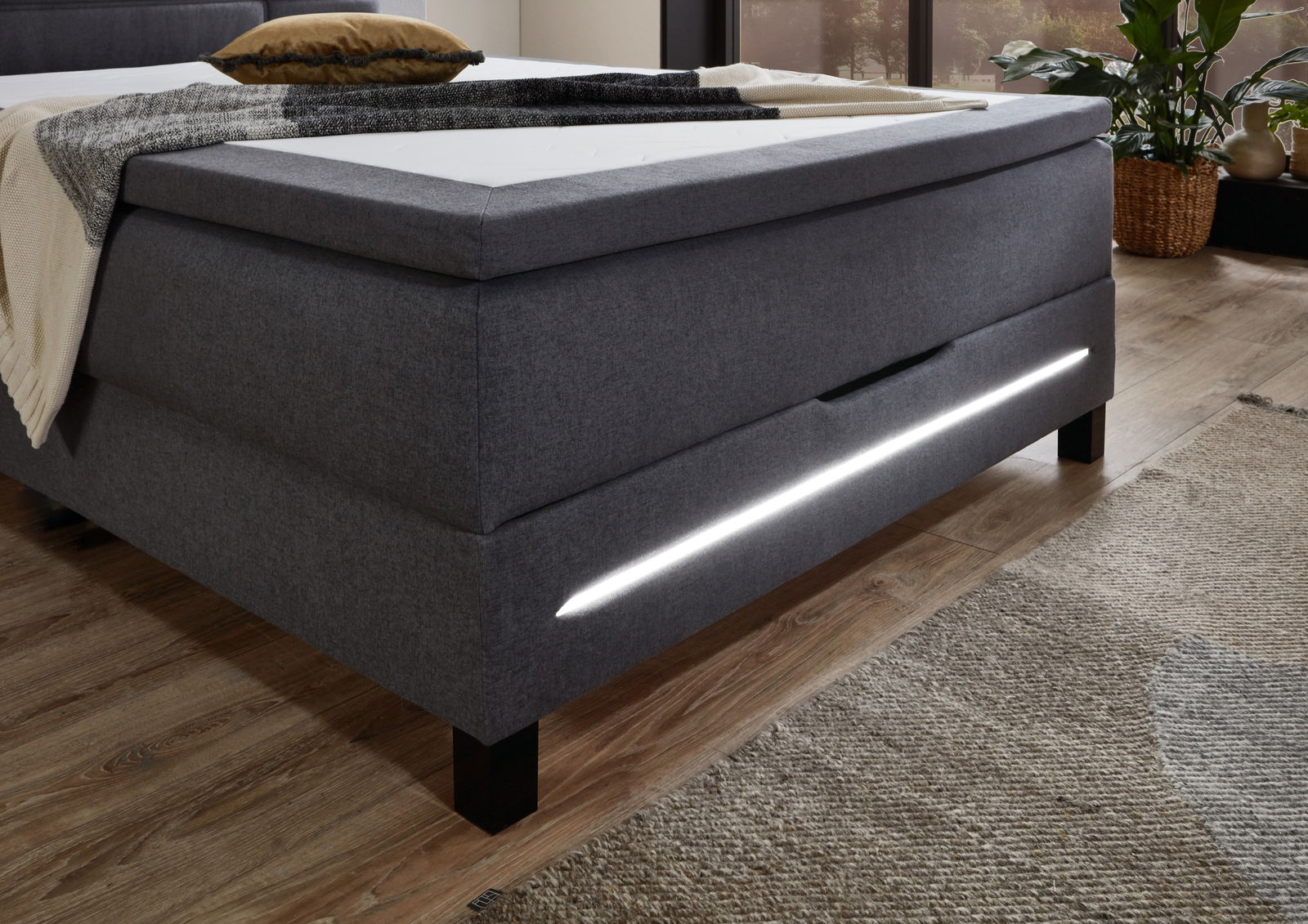 Seitliche Perspektive eines modernen grauen Boxspringbetts mit integrierter LED-Beleuchtung und stilvollem Design.