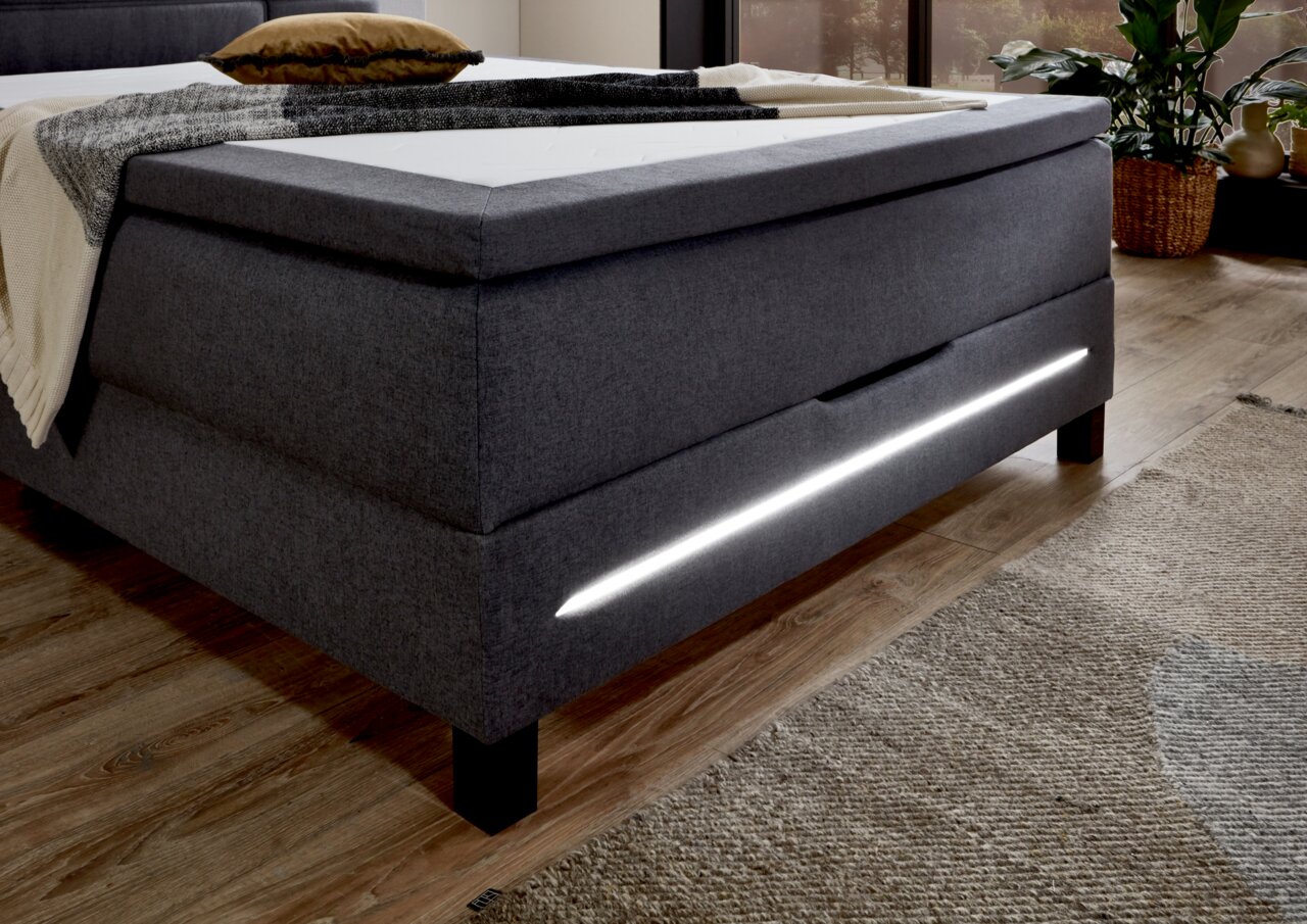 Seitliche Perspektive eines modernen grauen Boxspringbetts mit integrierter LED-Beleuchtung und stilvollem Design.