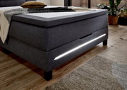Seitliche Perspektive eines modernen grauen Boxspringbetts mit integrierter LED-Beleuchtung und stilvollem Design.