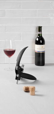Hebelkorkenzieher mit Folienschneider auf einem Tisch, im Hintergrund eine Flasche Rotwein und ein gefülltes Weinglas, seitliche Perspektive.
