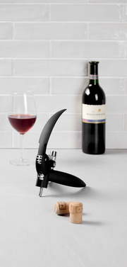 Hebelkorkenzieher mit Folienschneider auf einem Tisch, im Hintergrund eine Flasche Rotwein und ein gefülltes Weinglas, seitliche Perspektive.