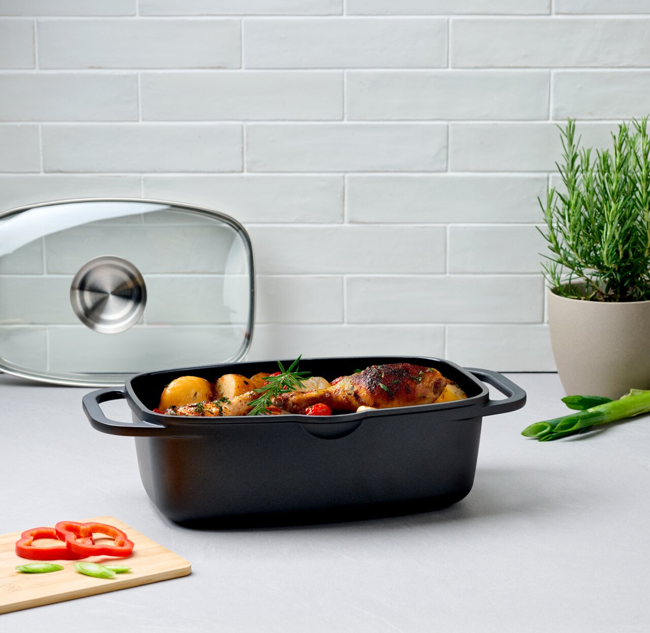 Quadratischer Alugussbräter mit Deckel, gefüllt mit gebratenem Hähnchen und Gemüse, auf einer Küchenarbeitsplatte. Perspektive von vorne.