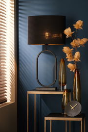 Moderner Lampenfuß JAMIRI in ovaler Form, schwarz, auf einem Tisch vor einer blauen Wand, seitliche Perspektive.