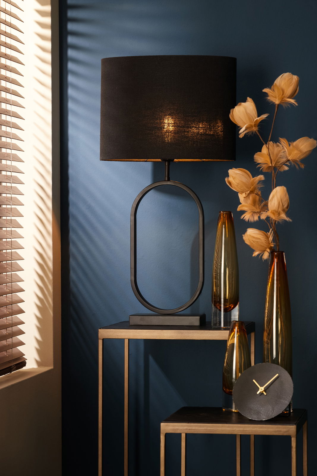 Moderner Lampenfuß JAMIRI in ovaler Form, schwarz, auf einem Tisch vor einer blauen Wand, seitliche Perspektive.