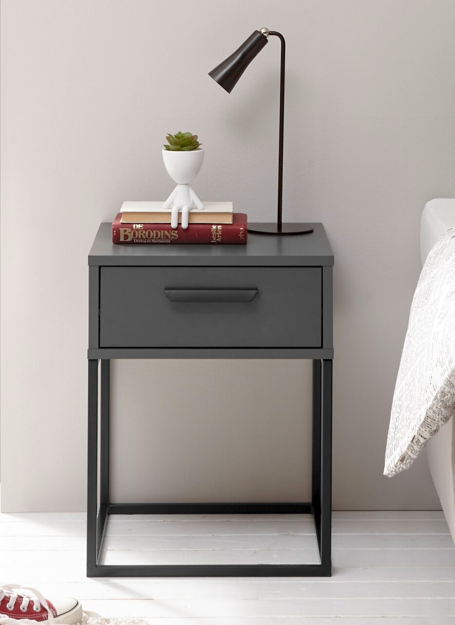 Moderne Nachtkommode in dunklem Grau mit einer Schublade, dekoriert mit einer Tischlampe, Büchern und einem kleinen Pflanzgefäß, seitliche Perspektive.