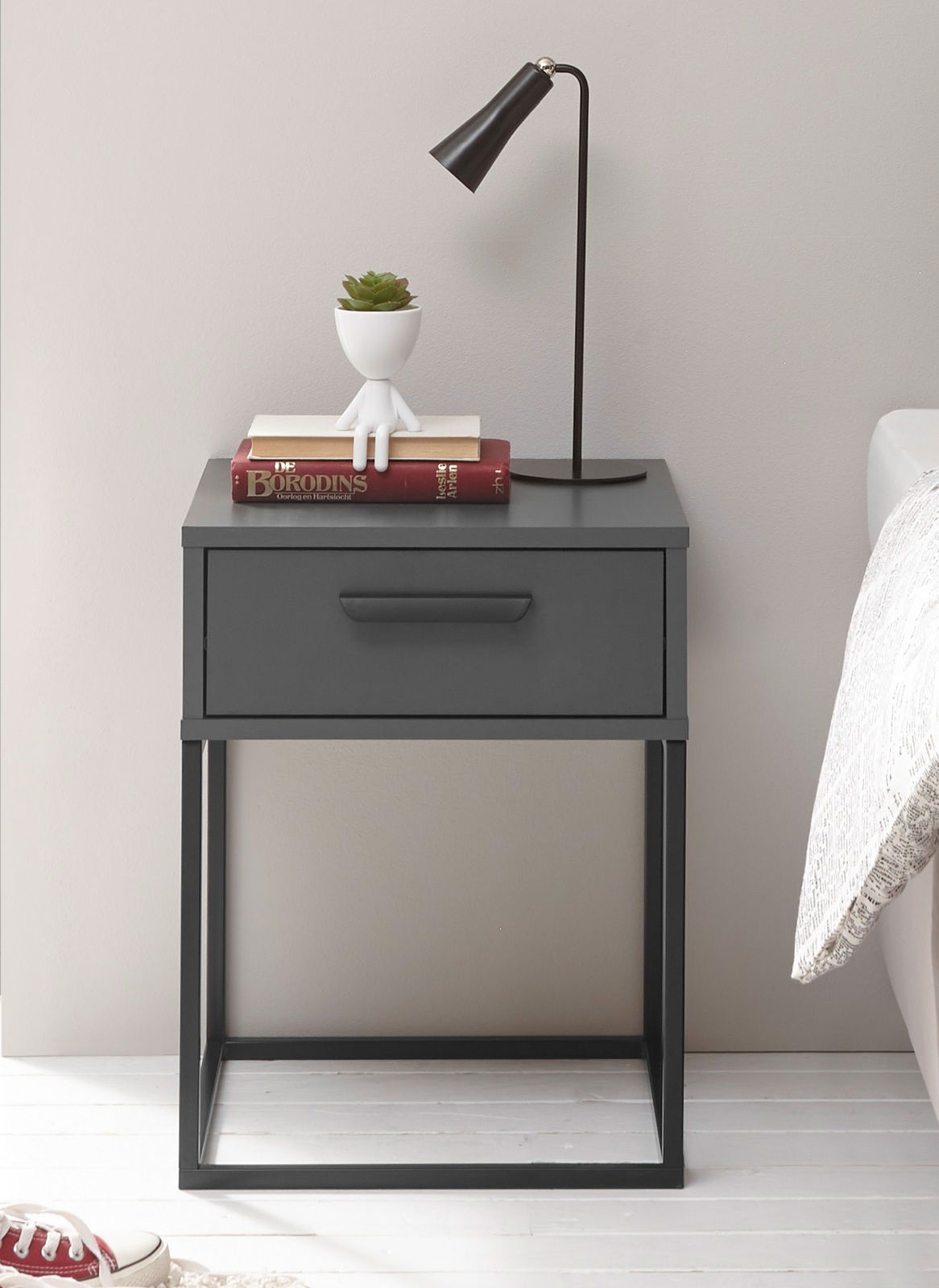 Moderne Nachtkommode in dunklem Grau mit einer Schublade, dekoriert mit einer Tischlampe, Büchern und einem kleinen Pflanzgefäß, seitliche Perspektive.