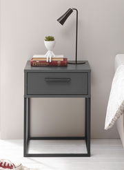 Moderne Nachtkommode in dunklem Grau mit einer Schublade, dekoriert mit einer Tischlampe, Büchern und einem kleinen Pflanzgefäß, seitliche Perspektive.