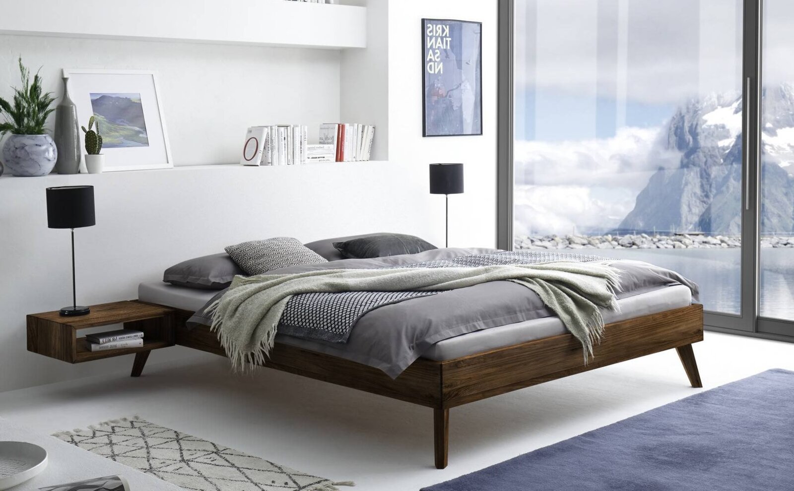 Modern houten tweepersoonsbed met grijs beddengoed en een plaid in een lichte slaapkamer, zijaanzicht vanuit een iets hoger standpunt