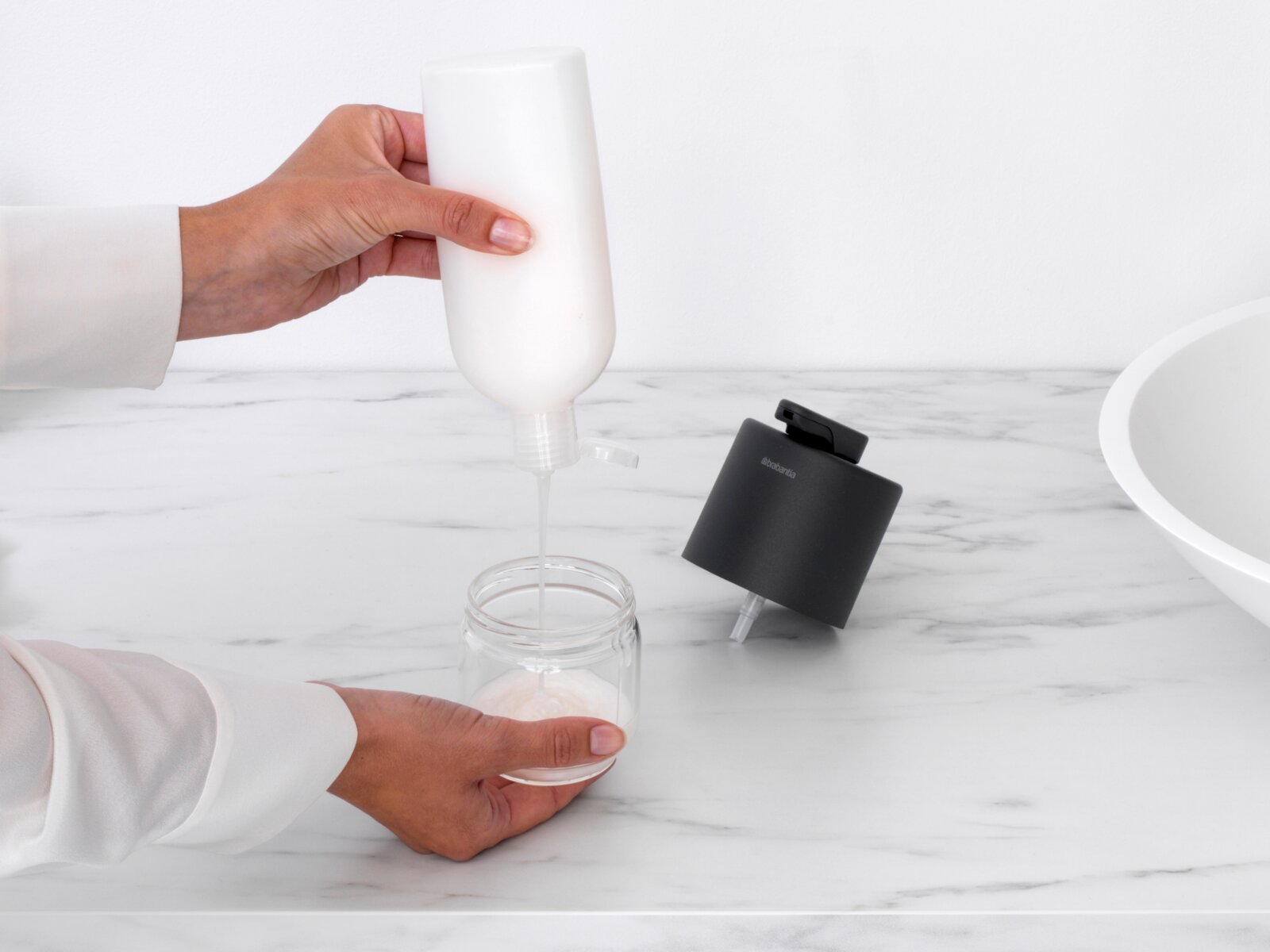 Zeepdispenser op een marmeren oppervlak, met een hand die zeep in een glazen pot giet. Aanzicht van bovenaf.