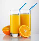 Zwei hohe Gläser mit Orangensaft und blauen Strohhalmen, umgeben von frischen Orangenhälften, aus der Frontalperspektive.