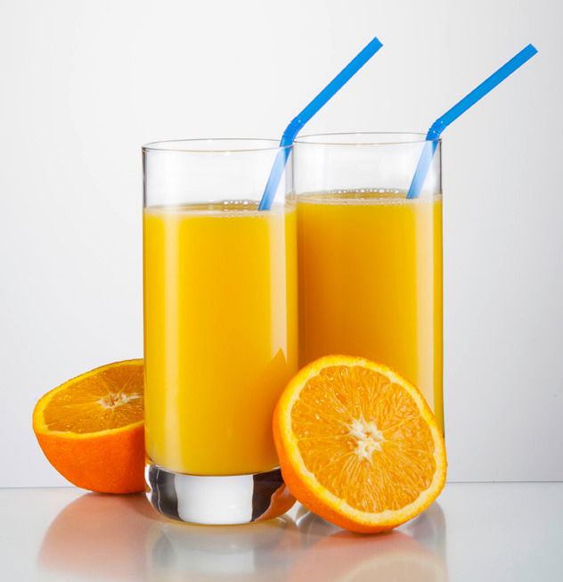 Zwei hohe Gläser mit Orangensaft und blauen Strohhalmen, umgeben von frischen Orangenhälften, aus der Frontalperspektive.