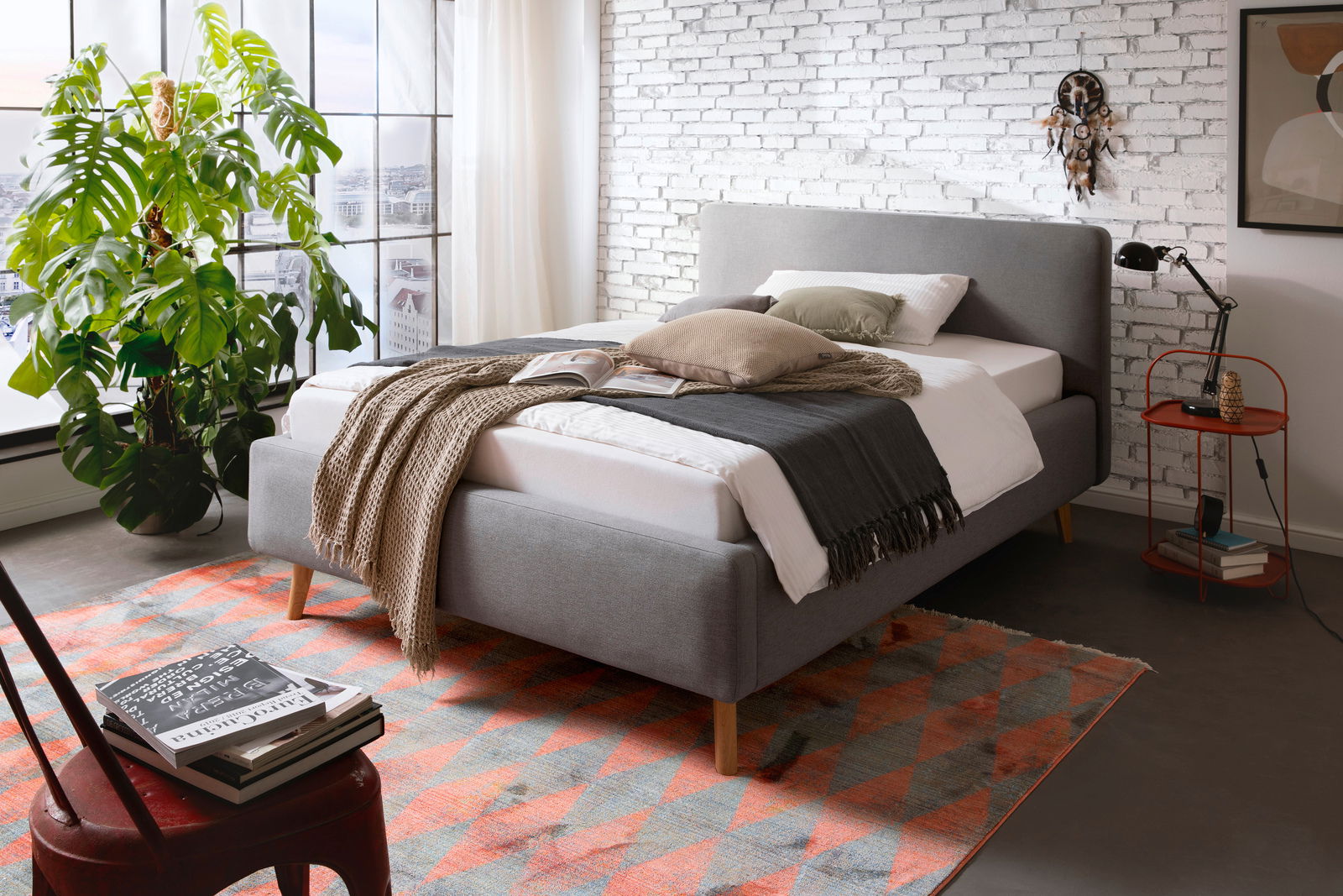 Z2 Polsterbett MATTIS Graues Polsterbett in modernem Schlafzimmer mit Blick von der Seite, dekoriert mit Kissen und Decken, neben einem großen Fenster und einer Pflanze.