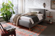 Grijs gestoffeerd bed in een moderne slaapkamer, gezien vanaf de zijkant, versierd met kussens en dekens, naast een groot raam en een plant.