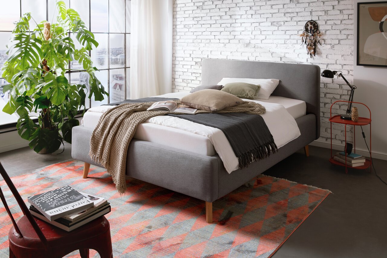 Grijs gestoffeerd bed in een moderne slaapkamer, gezien vanaf de zijkant, versierd met kussens en dekens, naast een groot raam en een plant.