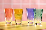 Vier bunte Trinkgläser in den Farben Orange, Gelb, Blau und Grün auf einem gelben Tisch vor einem rosa Hintergrund, aus der Vorderansicht fotografiert.