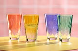 Vier bunte Trinkgläser in den Farben Orange, Gelb, Blau und Grün auf einem gelben Tisch vor einem rosa Hintergrund, aus der Vorderansicht fotografiert.