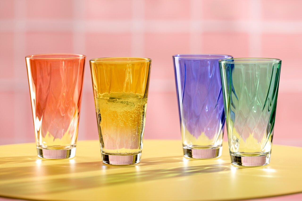 Vier bunte Trinkgläser in den Farben Orange, Gelb, Blau und Grün auf einem gelben Tisch vor einem rosa Hintergrund, aus der Vorderansicht fotografiert.