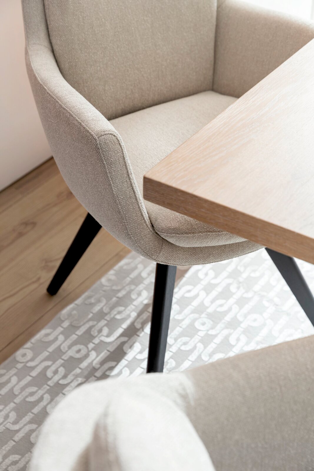 Beiger Armlehnstuhl mit schwarzem Gestell, seitliche Perspektive neben einem Holztisch auf einem gemusterten Teppich.