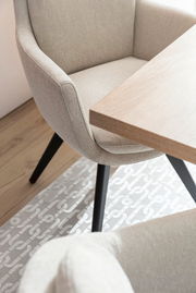 Beiger Armlehnstuhl mit schwarzem Gestell, seitliche Perspektive neben einem Holztisch auf einem gemusterten Teppich.