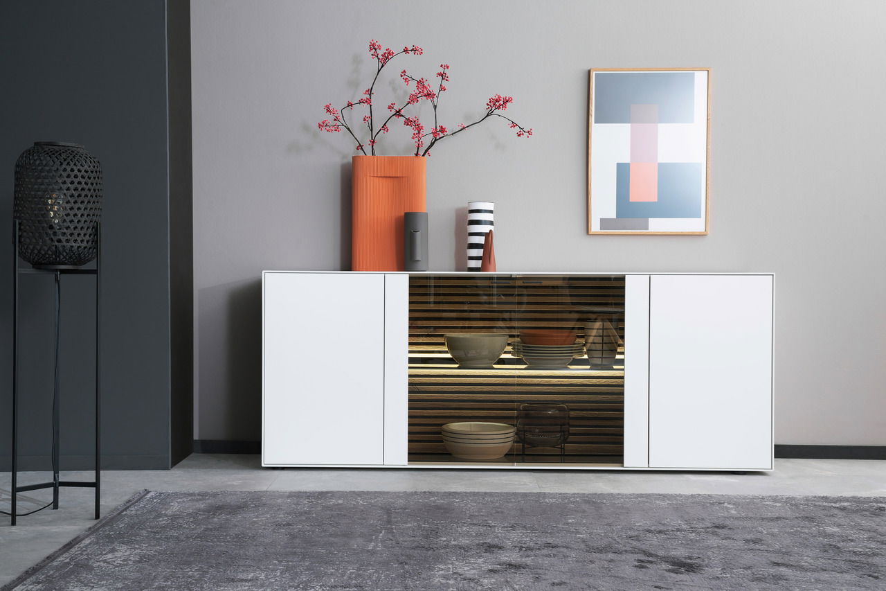 Modernes Sideboard in Weiß mit Glastür in der Mitte, dekoriert mit Vasen und Kunstwerk, aus frontaler Perspektive.