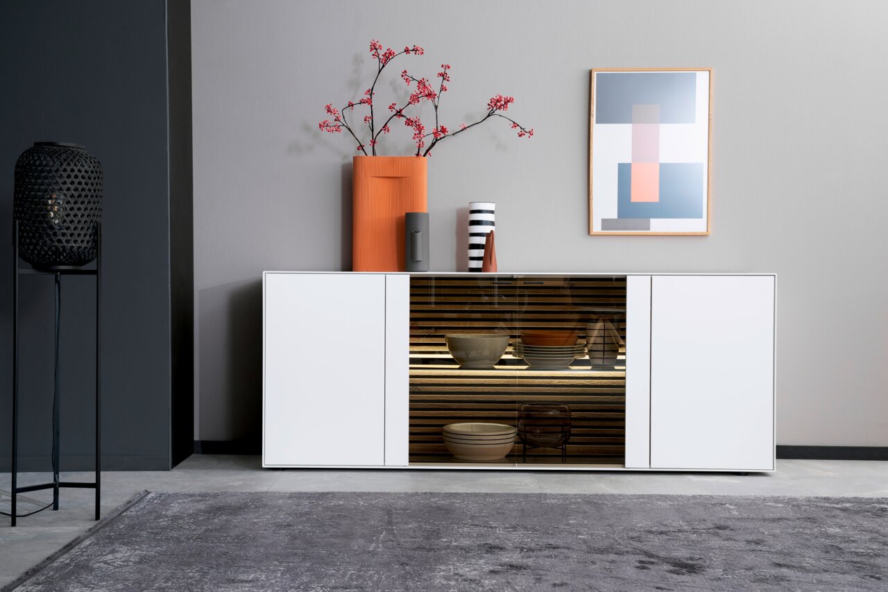 Modernes Sideboard in Weiß mit Glastür in der Mitte, dekoriert mit Vasen und Kunstwerk, aus frontaler Perspektive.