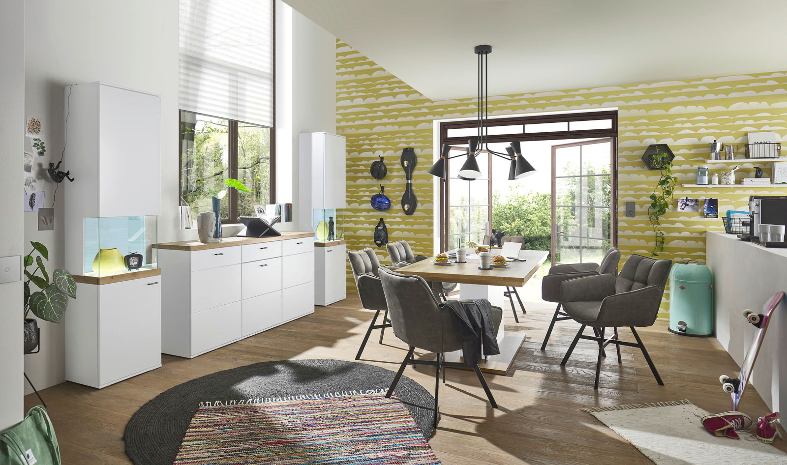 Moderne Esszimmer-Einrichtung mit weißem Sideboard und Vitrine, Esstisch mit grauen Stühlen, Blick aus der Perspektive des Raumes mit großen Fenstern und grüner Tapete.