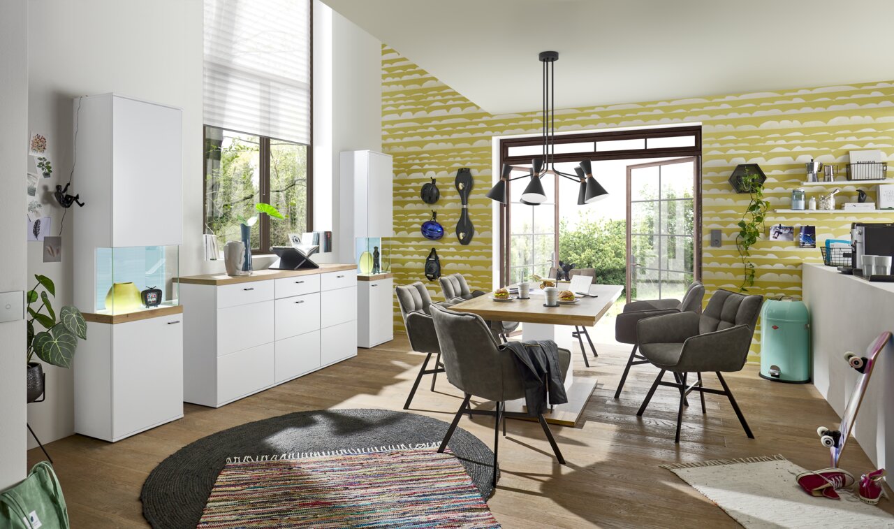 Set one by Musterring Sideboard SET ONE LANCASTER Moderne Esszimmer-Einrichtung mit weißem Sideboard und Vitrine, Esstisch mit grauen Stühlen, Blick aus der Perspektive des Raumes mit großen Fenstern und grüner Tapete.