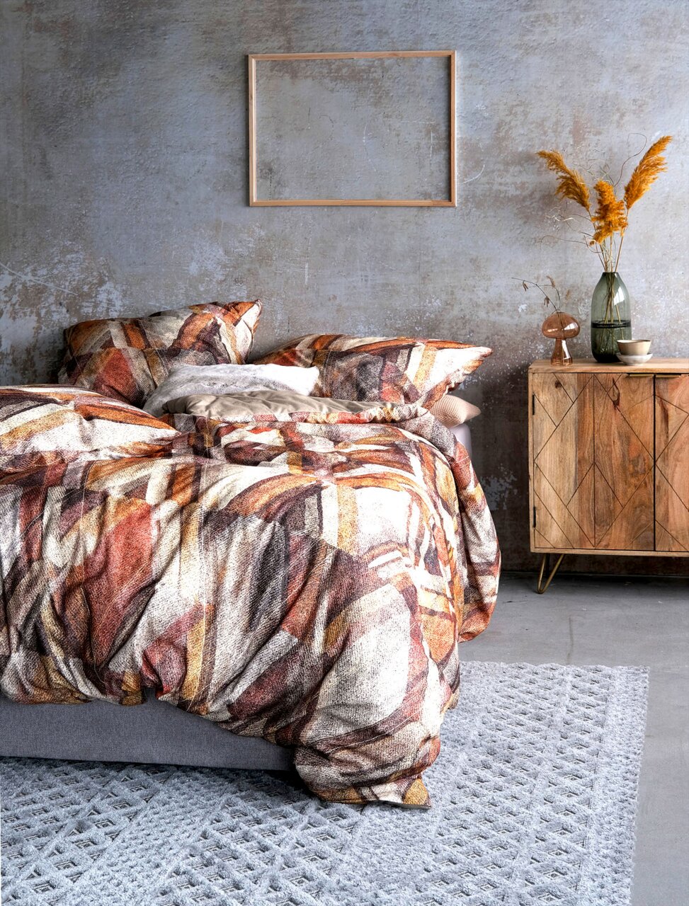 Bettwäsche-Set mit abstraktem Muster in warmen Erdtönen, auf einem grauen Bett in einem modern eingerichteten Raum mit Holzmöbeln und Dekorationen, aus seitlicher Perspektive fotografiert.