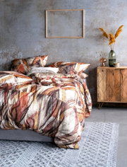 Bettwäsche-Set mit abstraktem Muster in warmen Erdtönen, auf einem grauen Bett in einem modern eingerichteten Raum mit Holzmöbeln und Dekorationen, aus seitlicher Perspektive fotografiert.