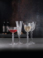 Vier elegante Kristallgläser, darunter ein Weißweinkelch, auf einer Tischoberfläche mit dunklem Hintergrund, seitliche Perspektive.