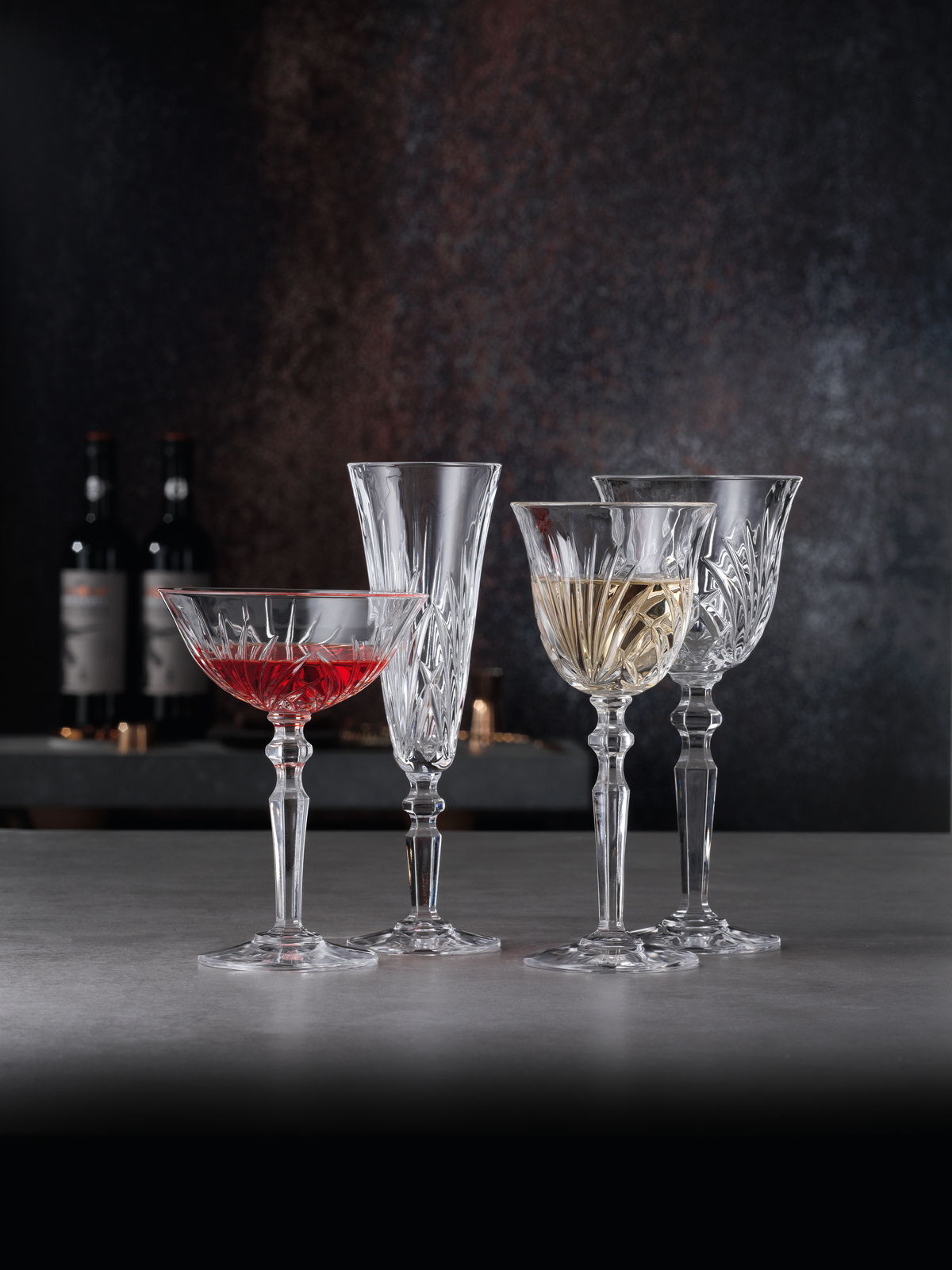 Vier elegante Kristallgläser, darunter ein Weißweinkelch, auf einer Tischoberfläche mit dunklem Hintergrund, seitliche Perspektive.