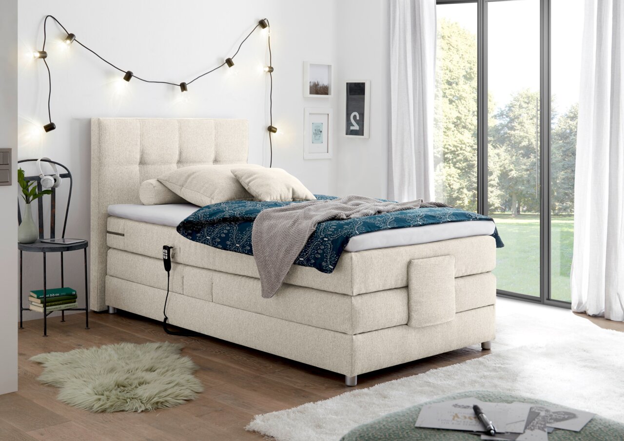Esposa Boxspringbett MANOLO Boxspringbett in einem modernen Schlafzimmer, seitliche Perspektive, mit hellem Stoffbezug, dekoriert mit Kissen und Decke, neben einem großen Fenster.