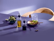 Zwei transparente Glasflaschen mit Ausgießer für Öl und Essig auf violettem Tisch, daneben Artischocke, Salatschüssel mit Holzlöffeln, Wasserglas und Holzstuhl im Hintergrund; Perspektive: leicht erhöhte seitliche Ansicht