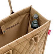 Nahaufnahme einer beigen Einkaufstasche mit Rautenmuster, die eine Flasche in einem Seitenfach zeigt. Perspektive von oben.