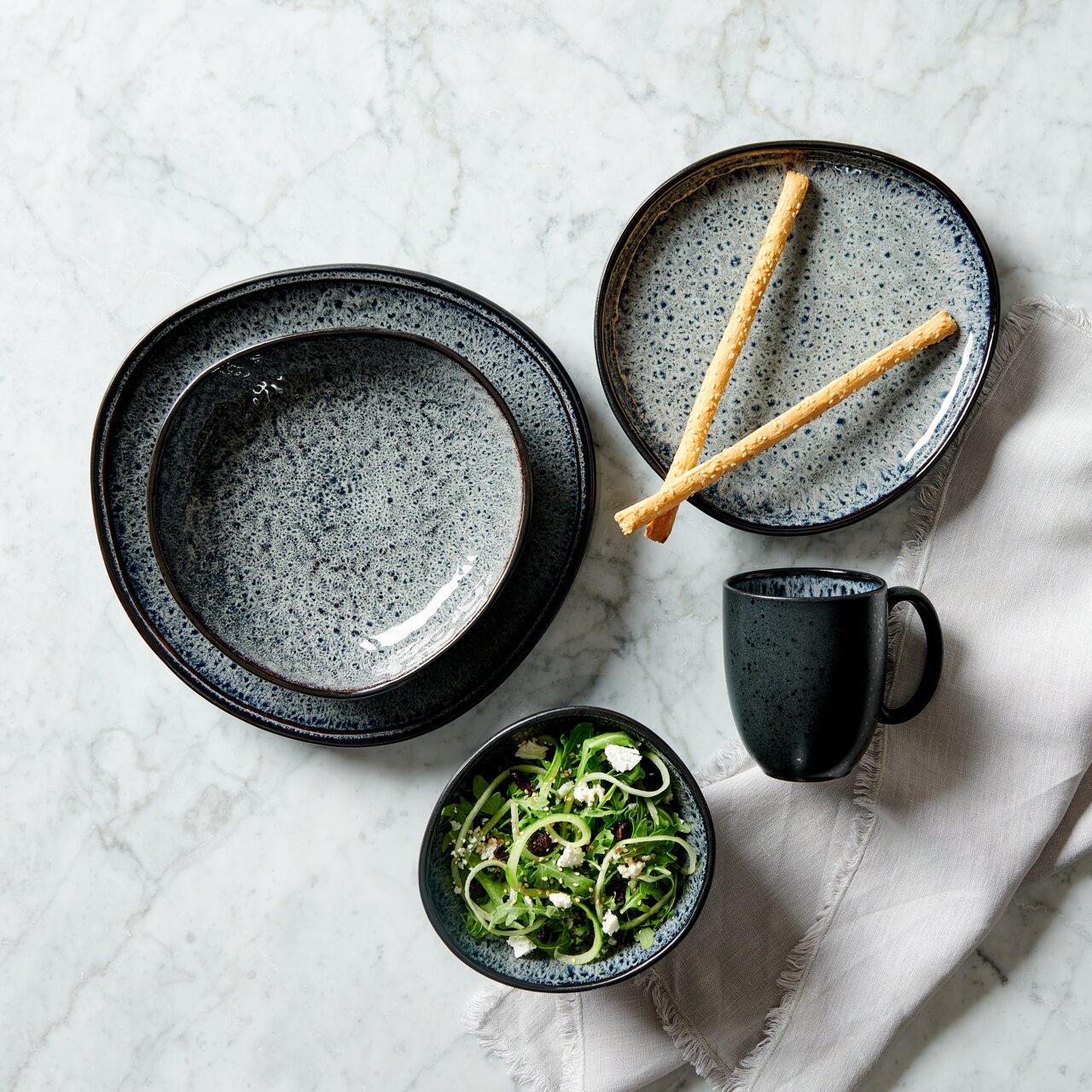 Vogelperspektive auf ein stilvolles, graues Kombiservice-Set mit Tellern, Schale, Tasse und Besteck auf einem Marmortisch.