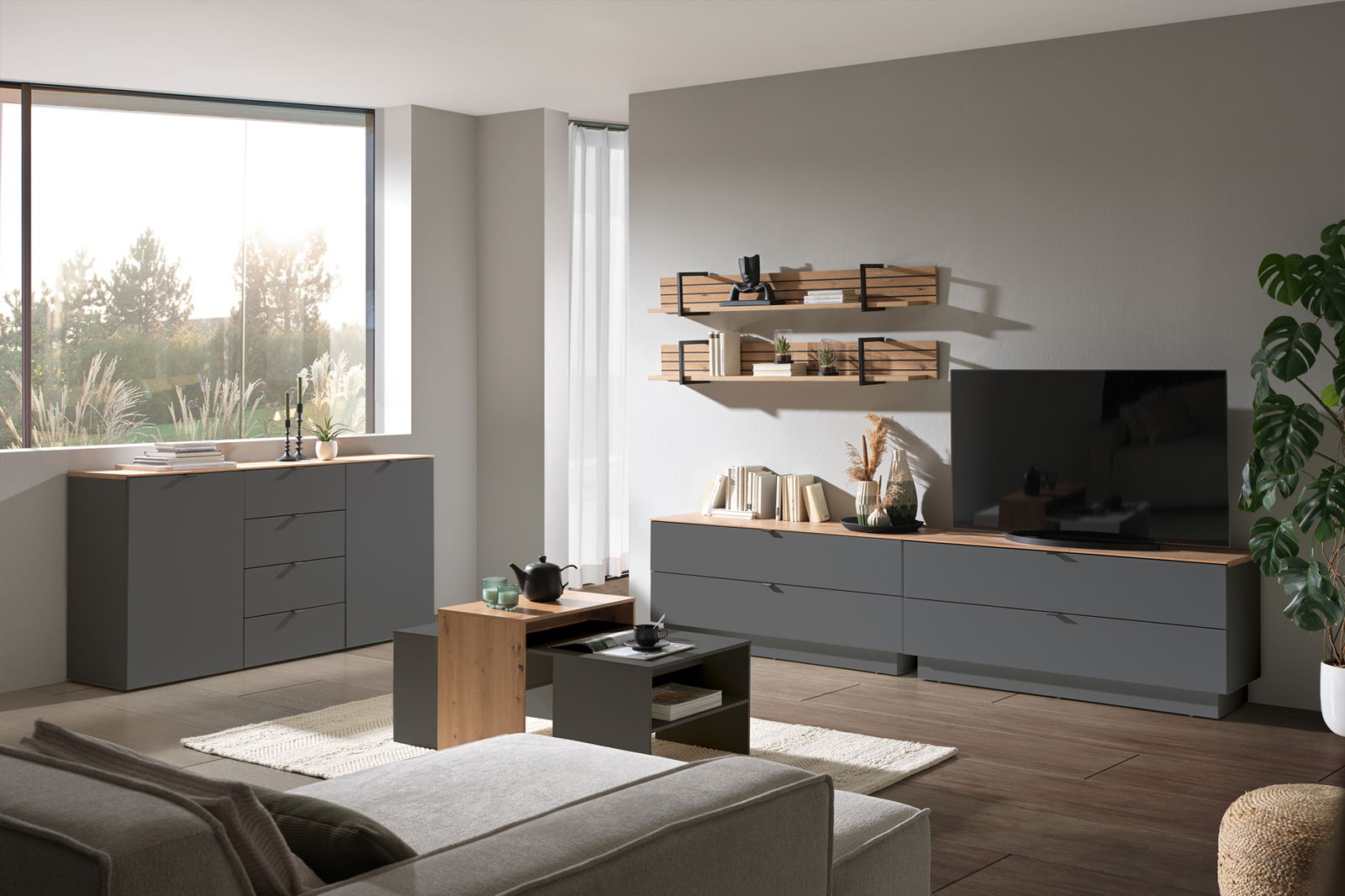 Moderne Wohnzimmermöbel in einem hellen Raum mit einem großen Fenster, bestehend aus einem grauen Sideboard, einem TV-Lowboard, einem Couchtisch und Wandregalen. Perspektive von vorne.