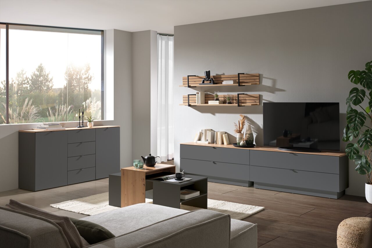 Moderne Wohnzimmermöbel in einem hellen Raum mit einem großen Fenster, bestehend aus einem grauen Sideboard, einem TV-Lowboard, einem Couchtisch und Wandregalen. Perspektive von vorne.