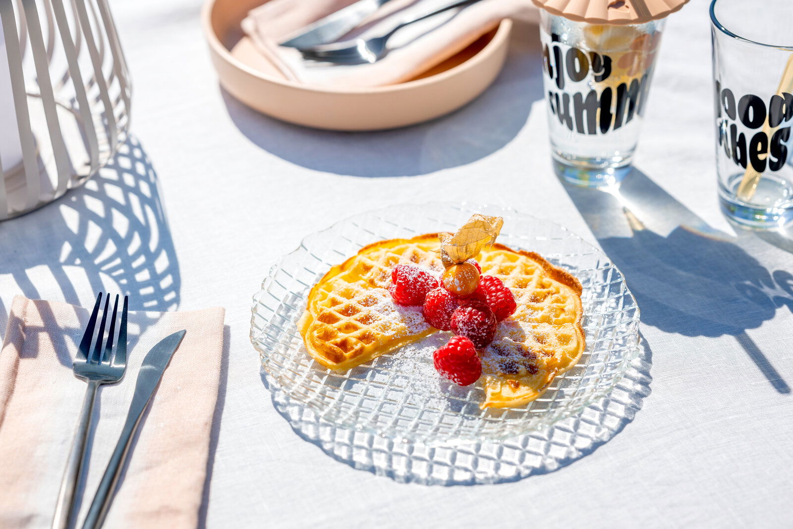 Ein gläserner Teller mit einer Waffel, garniert mit frischen Himbeeren und Puderzucker, auf einem Tisch im Sonnenlicht. Perspektive von oben.
