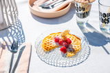 Ein gläserner Teller mit einer Waffel, garniert mit frischen Himbeeren und Puderzucker, auf einem Tisch im Sonnenlicht. Perspektive von oben.